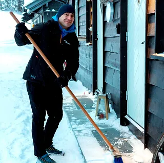 Mann fjerner snø med kost utenfor hytte